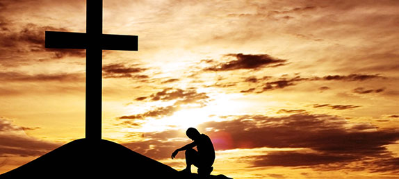 A person kneeling in front of a cross at sunset. Salvation can only be attained through Jesus' crucifixion and Resurrection.