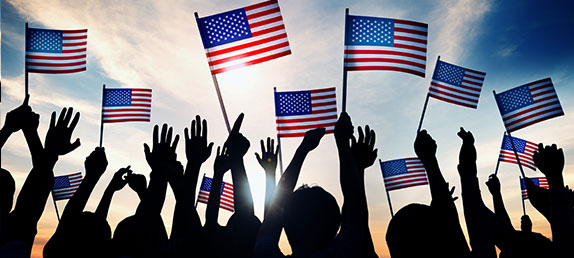 A group of people waving American flags silhouetted in the sun. Prayer makes America great and keeps God at the forefront.